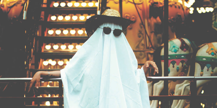 Adult in a white sheet ghost costume with a straw hat and sunglasses, posing against carnival lights for a playful Halloween photoshoot idea.