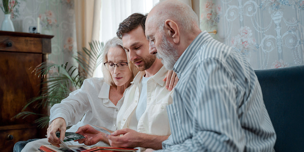 Adult son looking through an old family photo album with his aging parents before their home downsize.