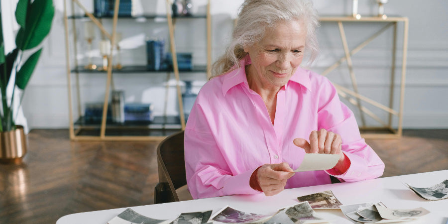 An older woman in a pink shirt sorting loose black-and-white photo prints spread across a table.