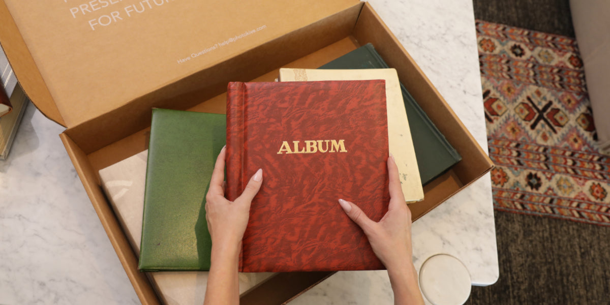 Hands placing a red photo album into a Photokive shipping box alongside other albums.