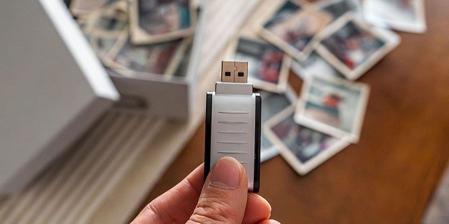 Hand holding a USB flash drive of digitized family photos in front of a box of loose vintage prints