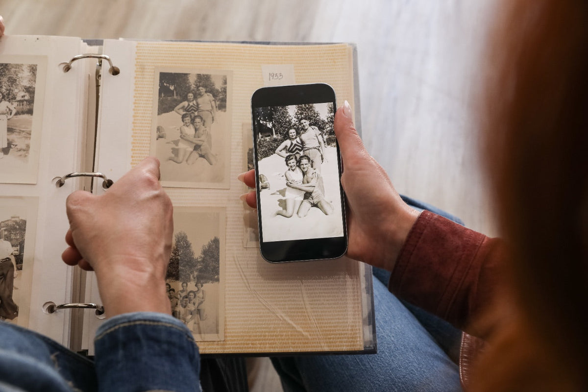 A woman comparing a restored family photo on her phone to the original in a vintage album